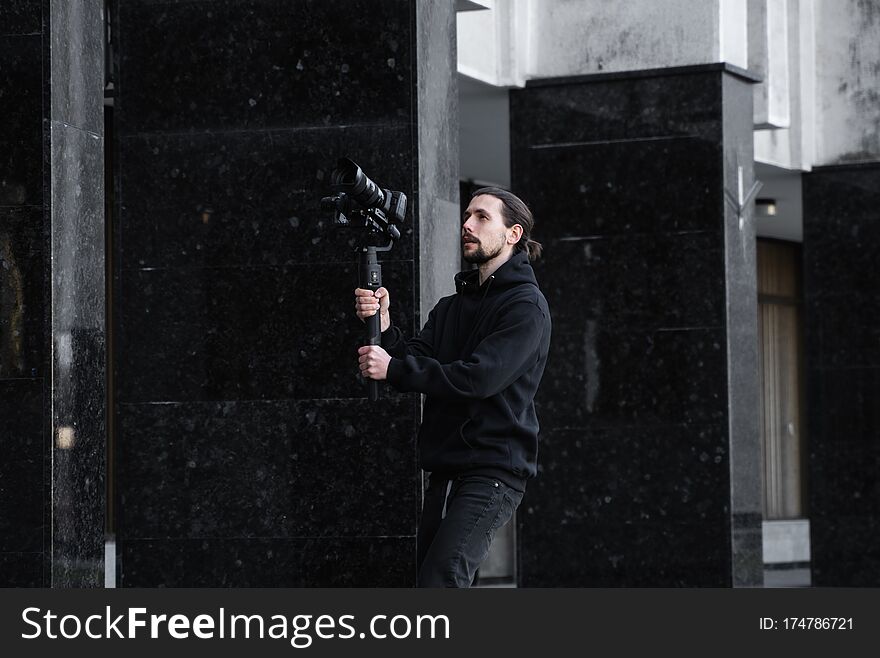 Young Professional videographer holding professional camera on 3-axis gimbal stabilizer. Pro equipment helps to make high quality video without shaking. Cameraman wearing black hoodie making a videos