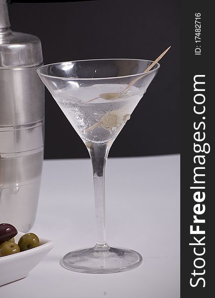 A glass of ice cold martini with cocktail shaker and olives in background