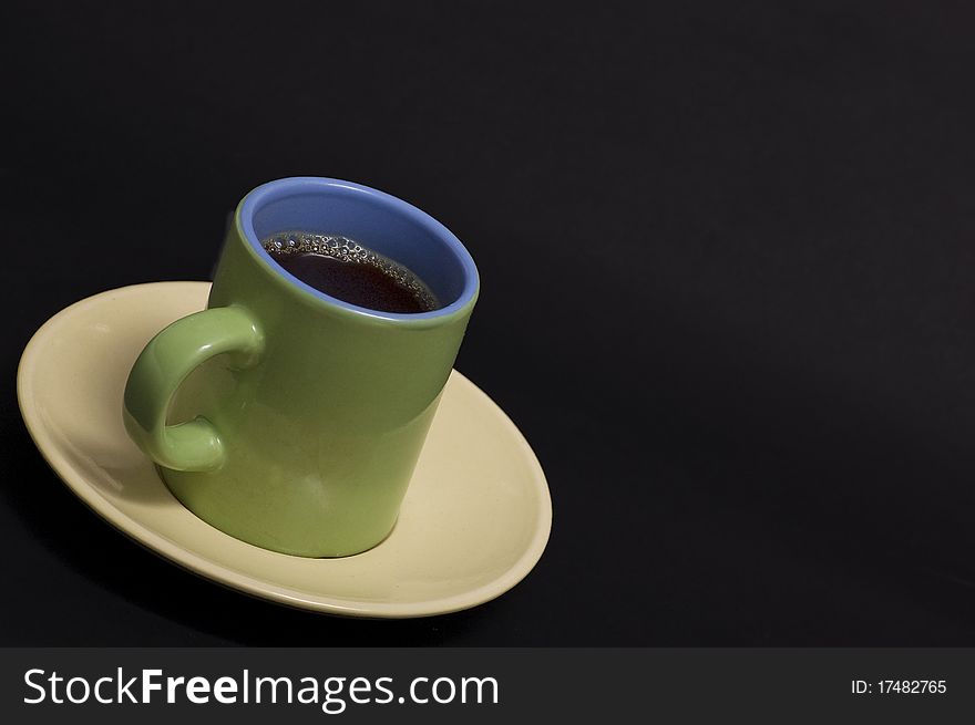 A green and blue cup of espresso on yellow saucer against black background. A green and blue cup of espresso on yellow saucer against black background