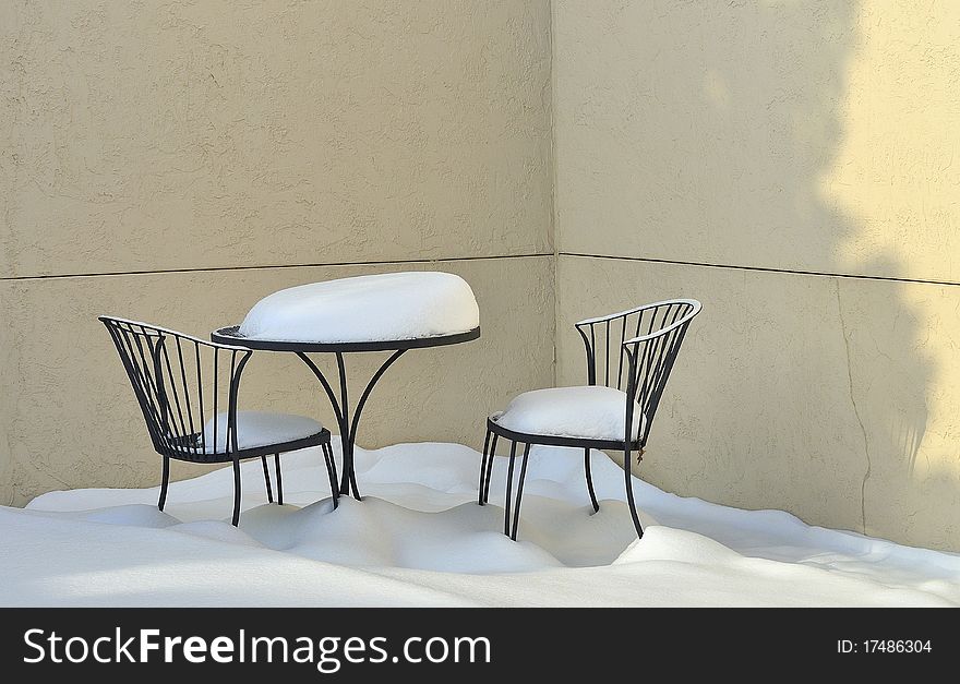 Patio table and two chairs outdoors on a empty patio with mounds of new fallen snow covering them and the floor. Patio table and two chairs outdoors on a empty patio with mounds of new fallen snow covering them and the floor.