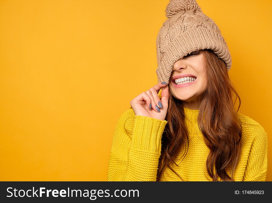 A brunette in a yellow sweater stands on a yellow background and smiles with white teeth, pulling the cap over her face. Horizontal photo