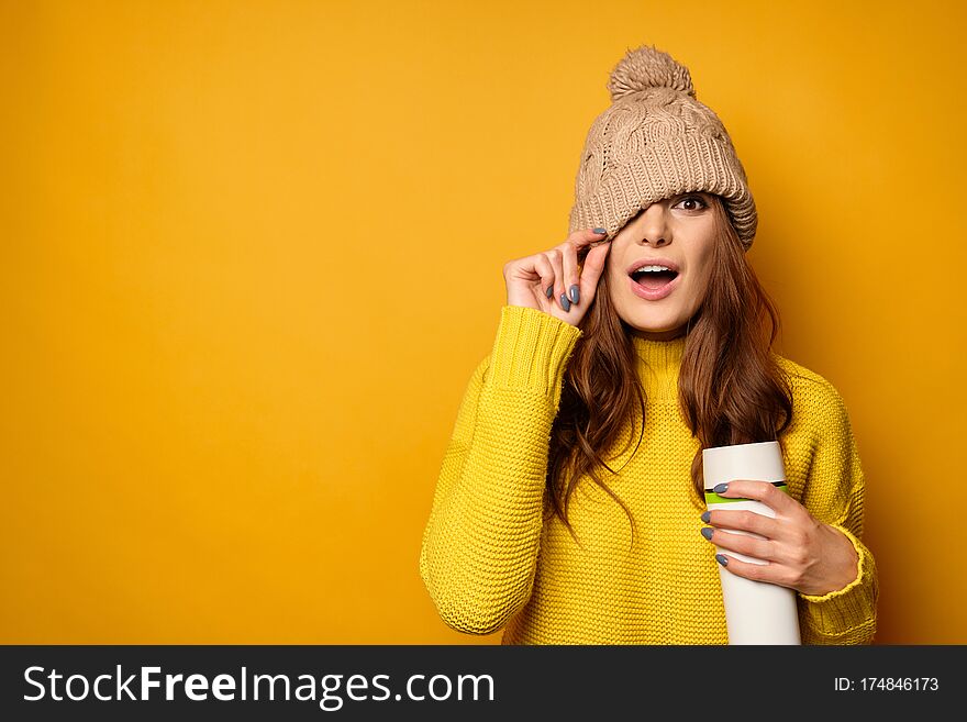 A brunette in a yellow sweater with a thermos is standing on a yellow background and pulling a hat over her face, her mouth open in surprise. Horizontal photo. A brunette in a yellow sweater with a thermos is standing on a yellow background and pulling a hat over her face, her mouth open in surprise. Horizontal photo