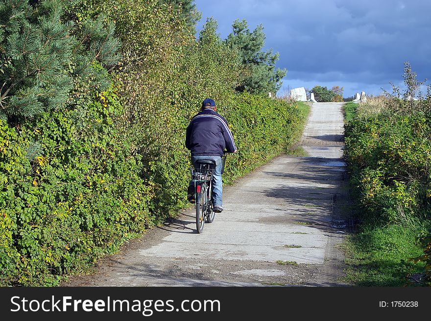 An older cyclist riding a bike, concrete path, green bushes. Wearing warm clothes.