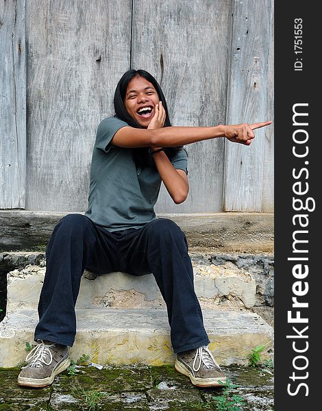 Asian tropical long-haired boy mockingly pointing with his finger in front of an old wooden weathered barn. Asian tropical long-haired boy mockingly pointing with his finger in front of an old wooden weathered barn.