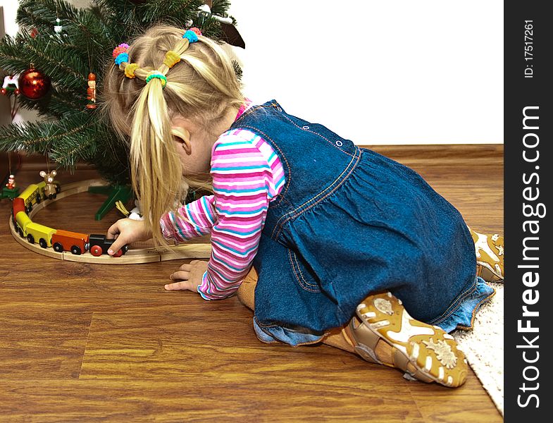 Blonde girl in jeans sarafan playing with toy wooden train, toys under the fir tree, copy space. Blonde girl in jeans sarafan playing with toy wooden train, toys under the fir tree, copy space