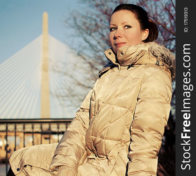 Girl in beige dawn parka sitting on a bench with a bridge on the background, Boston