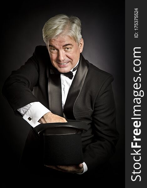 An elderly gray-haired prestidigitator in a black tuxedo with a black hat in his hands shows magic tricks on a black background. . An elderly gray-haired prestidigitator in a black tuxedo with a black hat in his hands shows magic tricks on a black background