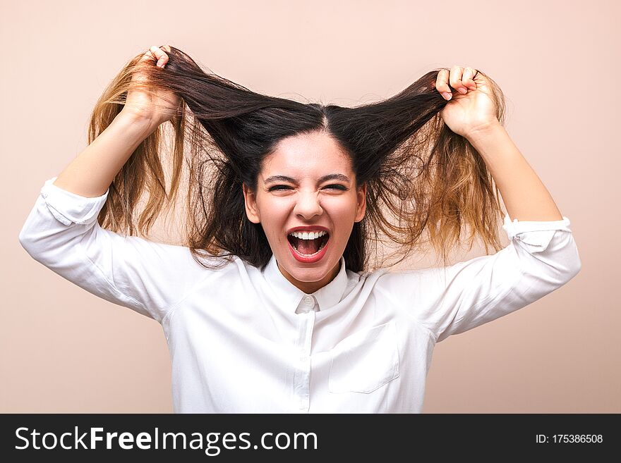 Attractive Brunette In White Shirt Pulls Her Hairs And Screams Against Pink Background. Woman Getting Crazy