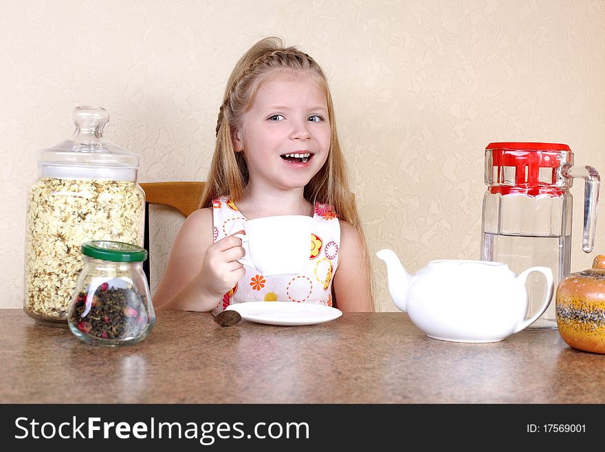 Little girlwith cup of hot drink, at table with tea, jasmin petals and teapot against yellow wall in home. Little girlwith cup of hot drink, at table with tea, jasmin petals and teapot against yellow wall in home