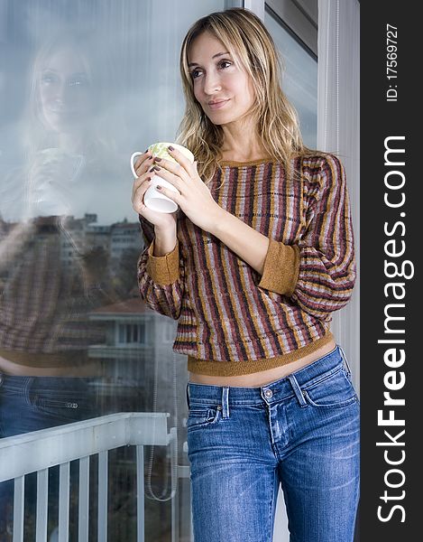 Blond woman holding a beverage mug and looking through her window. Blond woman holding a beverage mug and looking through her window