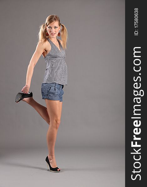 young blonde fashion model girl standing on one leg holding next foot. Model wearing short denim shorts, check top and stilettos. young blonde fashion model girl standing on one leg holding next foot. Model wearing short denim shorts, check top and stilettos.