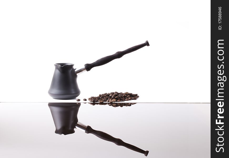 Coffee maker from dark metal with the long wooden handle of dark color and coffee grains fried are reflected in a white background from a table smooth surface. Coffee maker from dark metal with the long wooden handle of dark color and coffee grains fried are reflected in a white background from a table smooth surface