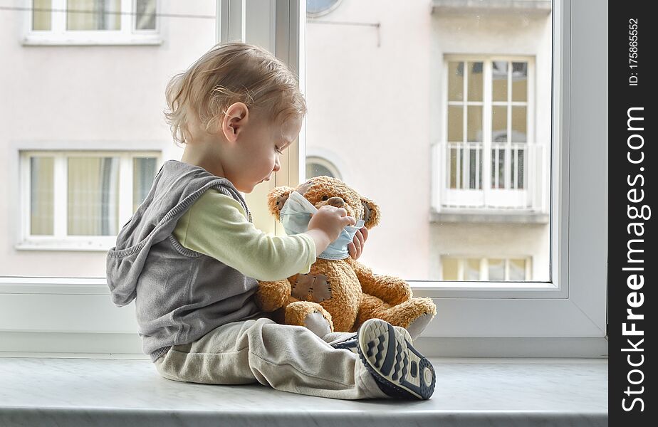 Child In Home Quarantine Playing At The Window With His Sick Teddy Bear Wearing A Medical Mask Against Viruses During Coronavirus