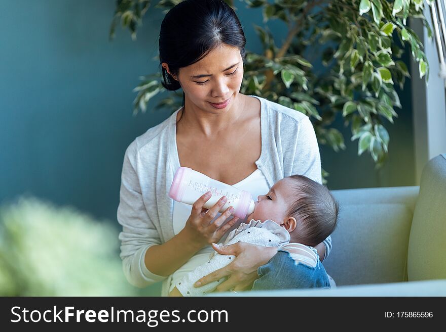 Shot of happy young mother feeding her baby daughter with feeding bottle while sitting on sofa at home