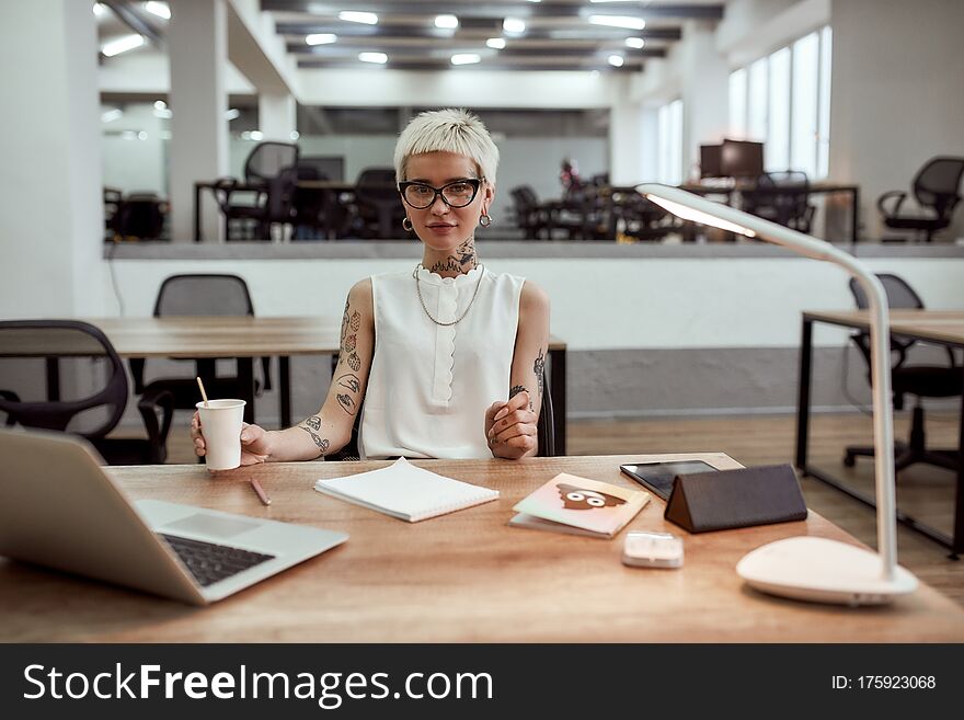 Little Break. Young Tattooed Business Woman In Eyewear Holding Cup Of Coffee And Looking At Camera While Working Alone