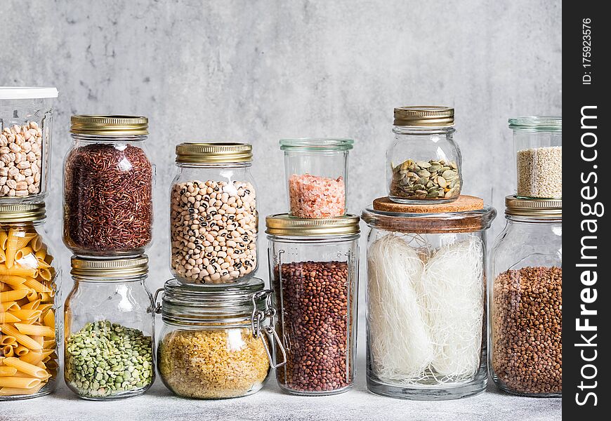 Glass jars with various cereals and seeds - peas split, sunflower and pumpkin seeds, beans, rice, pasta, oatmeal, lentils, bulgur on grey background