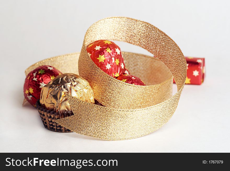 Chocolate golden sphere lay near a hill of sweets in a red metallic paper with golden stars on white background. Decorate of composition is golden strip. Chocolate golden sphere lay near a hill of sweets in a red metallic paper with golden stars on white background. Decorate of composition is golden strip.