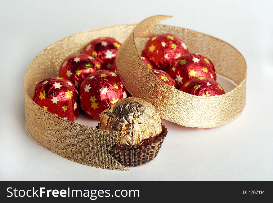 Chocolate golden sphere lay near a group of sweets in a red metallic paper with golden stars in the form of heard on white background. Decorate of composition is golden strip. Chocolate golden sphere lay near a group of sweets in a red metallic paper with golden stars in the form of heard on white background. Decorate of composition is golden strip.