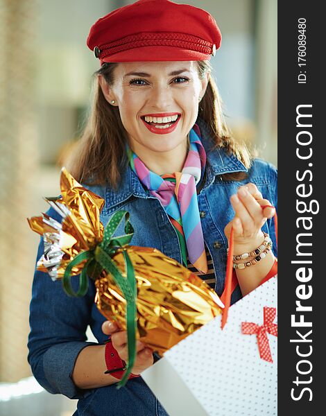 Happy young female in a jeans shirt and red hat at modern home in sunny spring day taking out wrapped in gold foil big easter egg from white polka dot shopping bag. Happy young female in a jeans shirt and red hat at modern home in sunny spring day taking out wrapped in gold foil big easter egg from white polka dot shopping bag