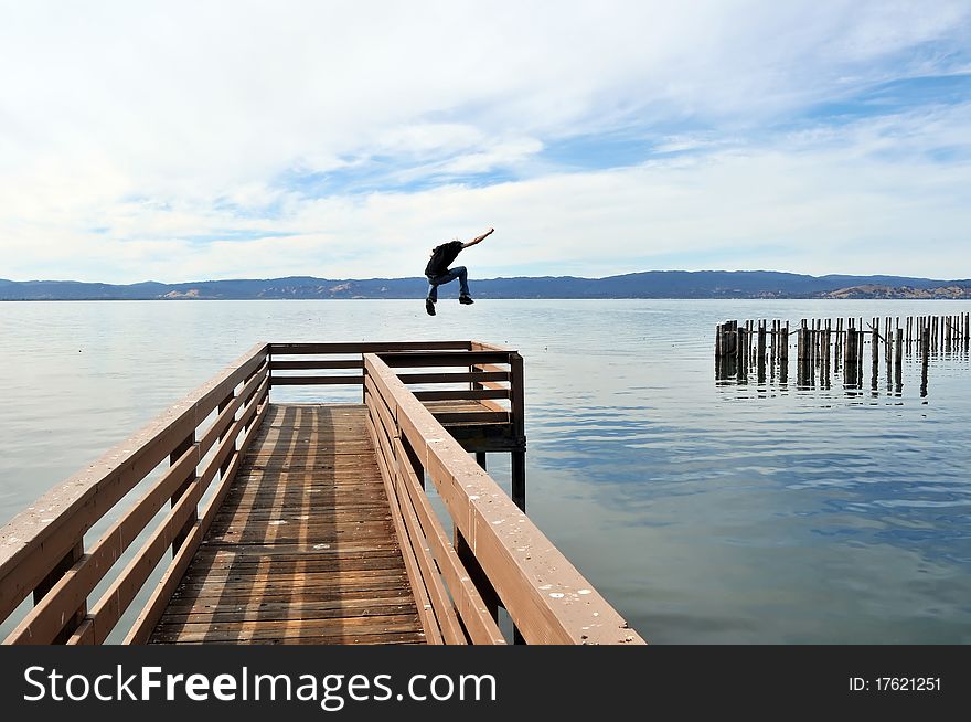 A man jumping a long narrow wooden pier into the calm blue ocean water with outstretched victorious arms. A man jumping a long narrow wooden pier into the calm blue ocean water with outstretched victorious arms.