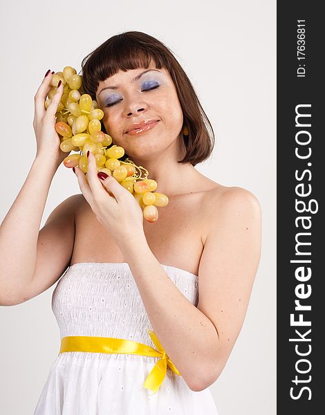 Beautiful girl in white dress with yellow tape is holding green grapes. She is isolated on a white background. Beautiful girl in white dress with yellow tape is holding green grapes. She is isolated on a white background.