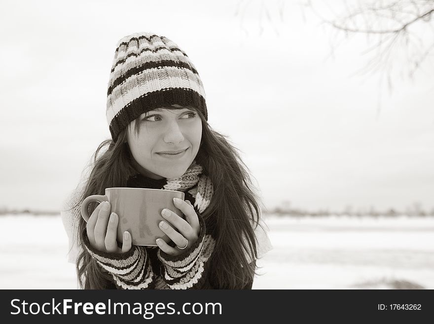 Young beautiful smiling woman in a striped hat and mittens sitting on the street with a large cup in his hand. Young beautiful smiling woman in a striped hat and mittens sitting on the street with a large cup in his hand