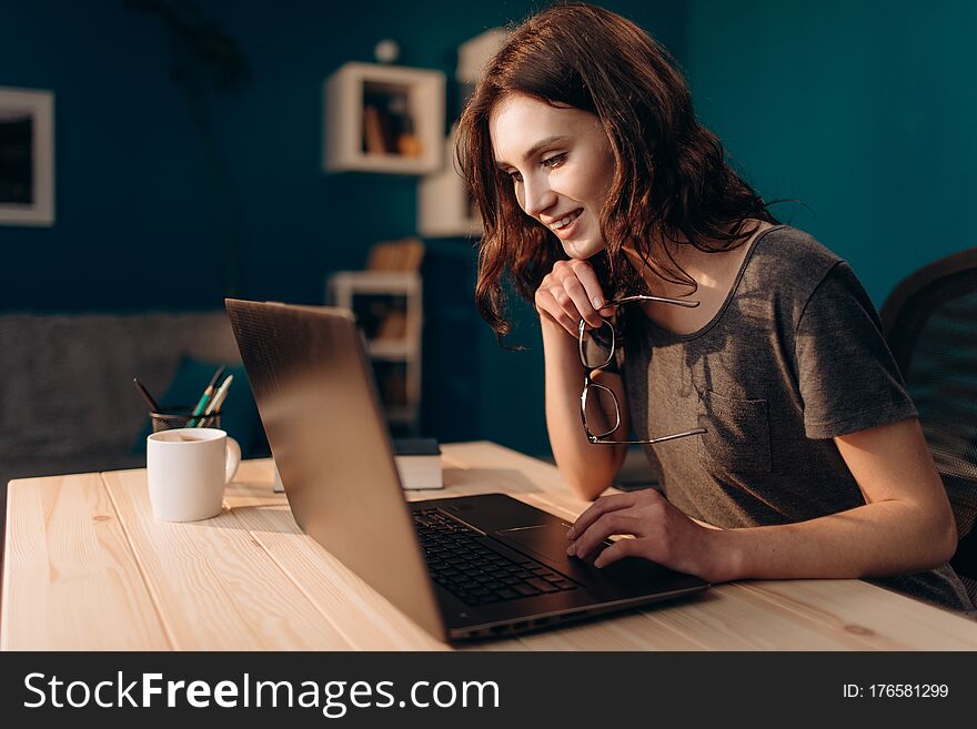 Cheerful woman with brown hair in casual outfit working on laptop at evening time. Young girl using digital gadget while sitting at table at home. Cheerful woman with brown hair in casual outfit working on laptop at evening time. Young girl using digital gadget while sitting at table at home.