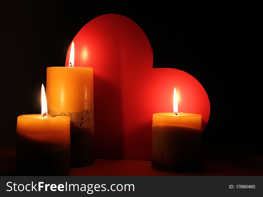 Lit Candles are illuminating a red love heart on a black background. Lit Candles are illuminating a red love heart on a black background