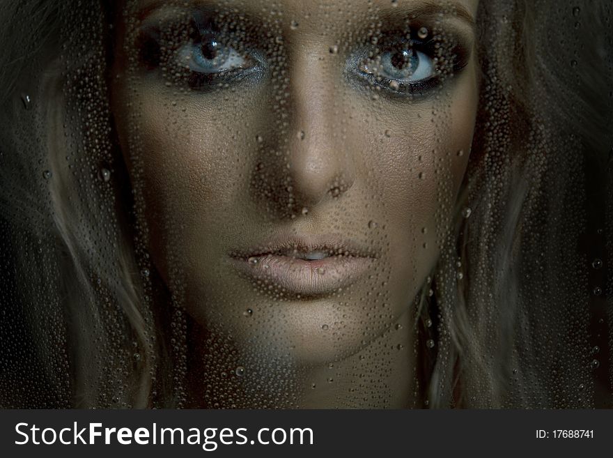 Face of gorgeous woman with a big blue eyes in the wet window. Face of gorgeous woman with a big blue eyes in the wet window