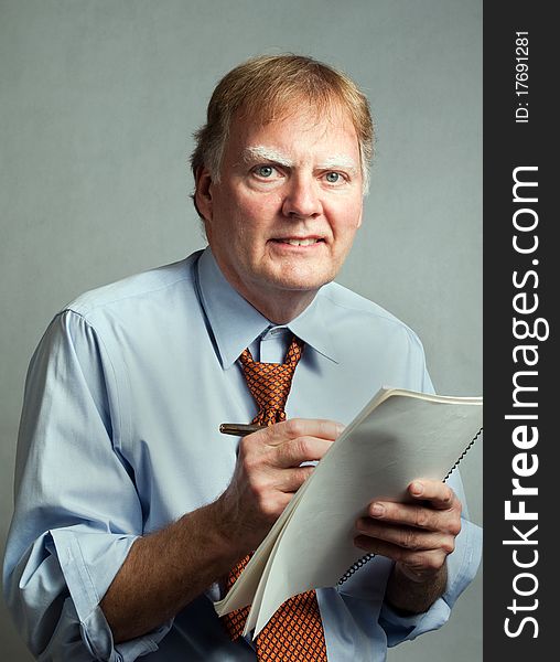 Businessman, professional, writing on paper, smiling and looking at the camera. The man is wearing a blue shirt and orange necktie and is in front of a white background. His shirt sleeves are rolled up and collar loosened. Businessman, professional, writing on paper, smiling and looking at the camera. The man is wearing a blue shirt and orange necktie and is in front of a white background. His shirt sleeves are rolled up and collar loosened.