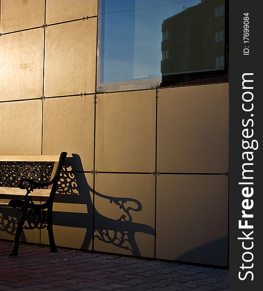 Bench and its shadow on a gold-yellow wall and a building reflection on the window