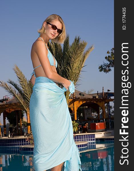 Picture of a blond beautiful smiling girl wearing blue pareo and standing near pool. Picture of a blond beautiful smiling girl wearing blue pareo and standing near pool