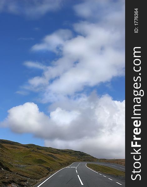 Empty down hill road in mountains with a dangerous right hand bend, with grass verges to either side, on a blue sky day with cumulus clouds. Located in the Brecon Beacons National Park, Wales, United Kingdom. Empty down hill road in mountains with a dangerous right hand bend, with grass verges to either side, on a blue sky day with cumulus clouds. Located in the Brecon Beacons National Park, Wales, United Kingdom.