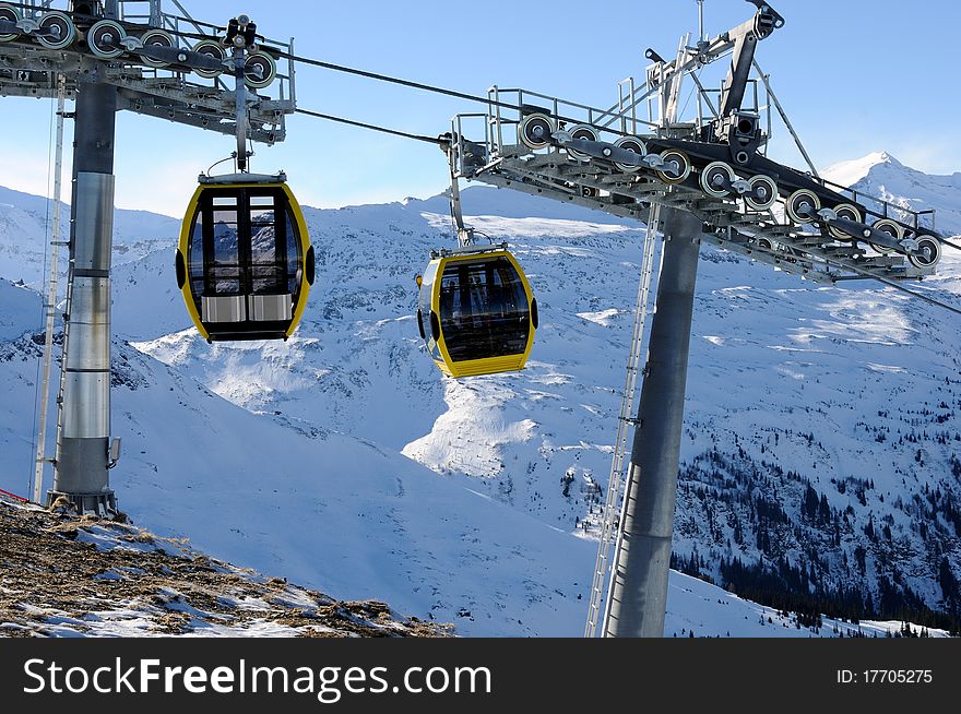 Two semitransparent yellow cabins on the cableway between steel supports with mountains. Two semitransparent yellow cabins on the cableway between steel supports with mountains