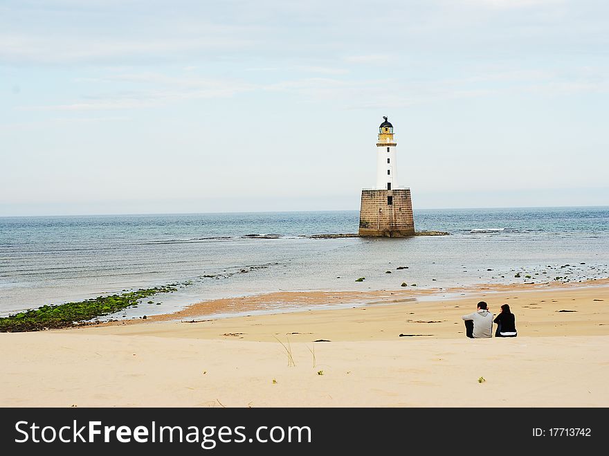Young couple siting on a beach with a white lighthouse. Young couple siting on a beach with a white lighthouse