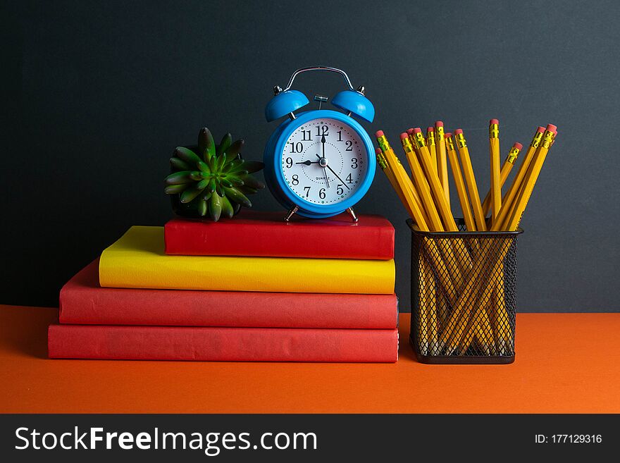 A stack of books, a green flower, blue clock and a glass of pencils stand on an orange background