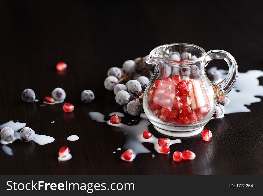 It is photo red granules of pomegranate in the small glass jug with bunch of dark grapes and drops of milk on a dark background. It is photo red granules of pomegranate in the small glass jug with bunch of dark grapes and drops of milk on a dark background.