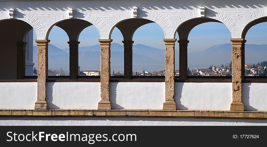 A view of the archways of the Alhambra fortress and palace in Granada, Spain with the nearby mountains and houses in the background. A view of the archways of the Alhambra fortress and palace in Granada, Spain with the nearby mountains and houses in the background.