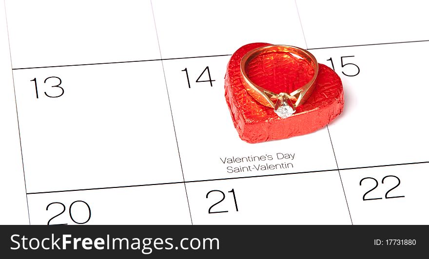 A angled side view of a red heart shaped chocolate candy with an engagement ring on top marking February 14th on the calendar. A angled side view of a red heart shaped chocolate candy with an engagement ring on top marking February 14th on the calendar.