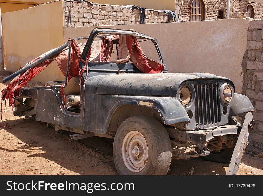 Old jeep without a bumper and without the rear wheels under a layer of dust and sand covered with the remains of the red carpet. Old jeep without a bumper and without the rear wheels under a layer of dust and sand covered with the remains of the red carpet.