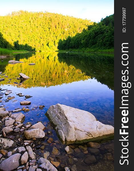 River in the evening. Bright sunlight reflected on a distant hill, covered with forests. Stones in the water in the foreground