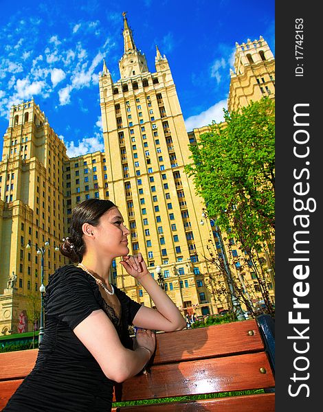 Beautiful Asian girl in black dress sitting on a bench on a lovely day on the background of high beautiful building. Beautiful Asian girl in black dress sitting on a bench on a lovely day on the background of high beautiful building