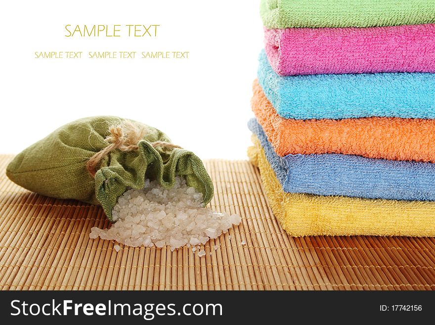 On a wooden mat lots of colorful bath towels stacked on each other lies near the fabric bag filled with bath salts. On a wooden mat lots of colorful bath towels stacked on each other lies near the fabric bag filled with bath salts.