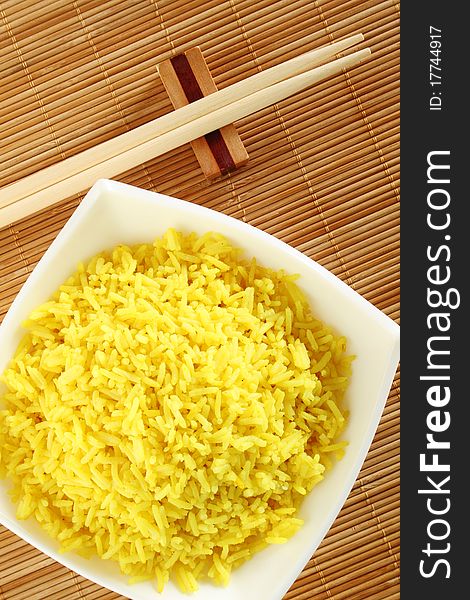 Boiled yellow rice and a white plate beside lie wooden sticks. Plate stands on a wooden mat. Boiled yellow rice and a white plate beside lie wooden sticks. Plate stands on a wooden mat
