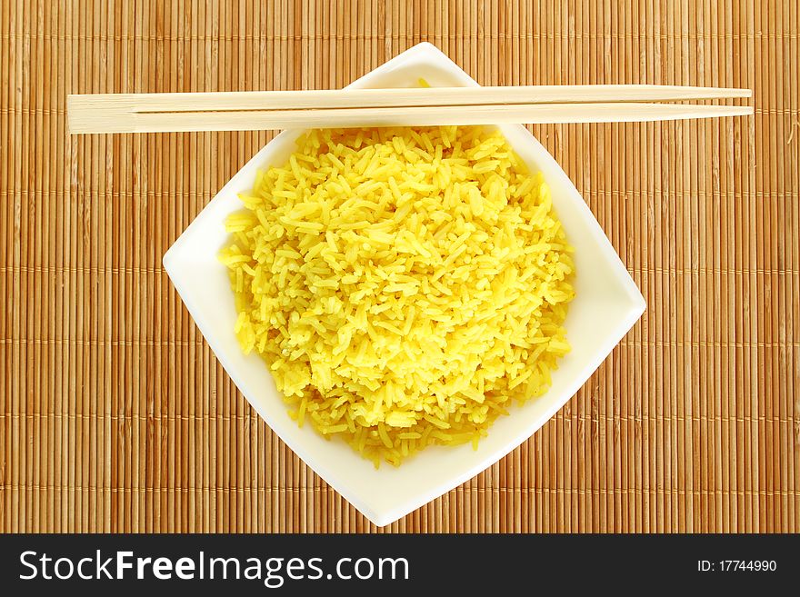 Boiled yellow rice and a white plate beside lie wooden sticks. Plate stands on a wooden mat. Boiled yellow rice and a white plate beside lie wooden sticks. Plate stands on a wooden mat