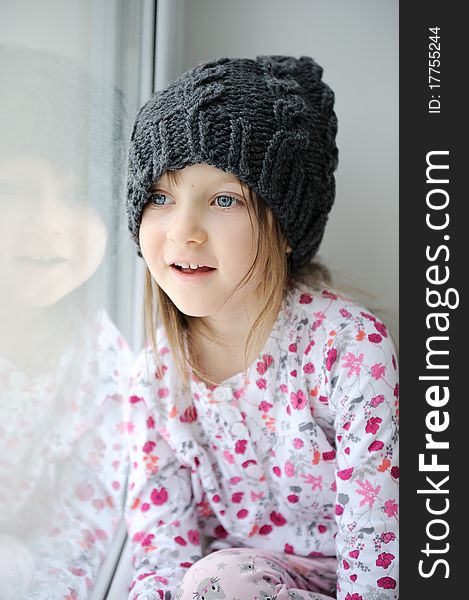 Adorable little kid girl with blue eyes in dark grey knit hat near the window. Adorable little kid girl with blue eyes in dark grey knit hat near the window