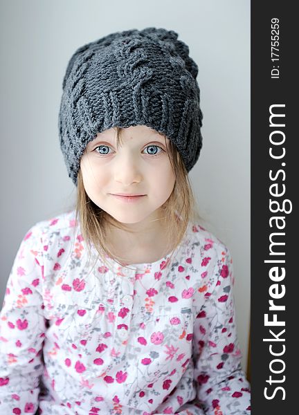 Adorable little kid girl with blue eyes in dark grey knit hat near the window looks into the camera. Adorable little kid girl with blue eyes in dark grey knit hat near the window looks into the camera