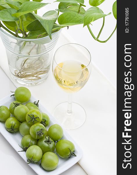 Glass of white wine with green plants and small green tomato on white background.