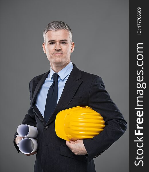 Architect or structural engineer carrying blueprints and hardhat under his arms standing smiling at the camera over grey with copy space. Architect or structural engineer carrying blueprints and hardhat under his arms standing smiling at the camera over grey with copy space