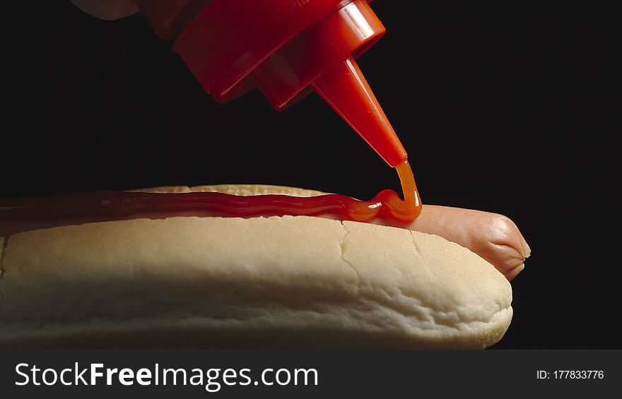 CLOSE UP: Ketchup bottle pouring tomato sause on a hot dog with sausage. Side view